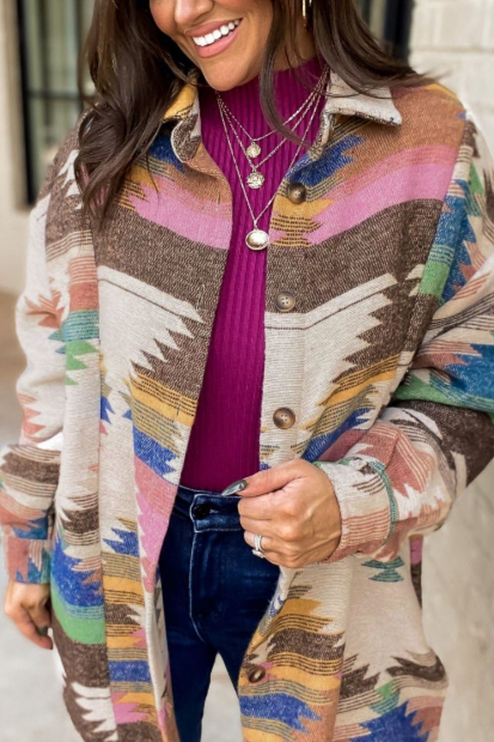 A woman smiling, wearing a multicolored geometric patterned long sleeve jacket with a V-neckline and button details, paired with blue jeans.