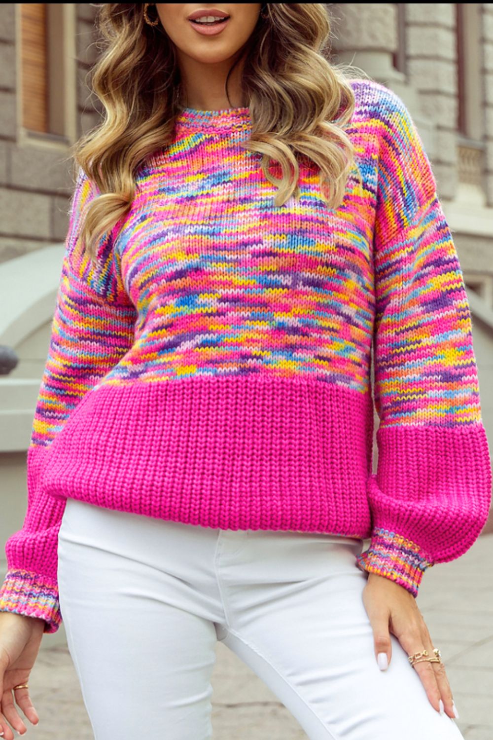 A woman wearing a multicolored, rainbow-striped confetti-patterned drop shoulder sweater in a vibrant pink hue, paired with white pants.